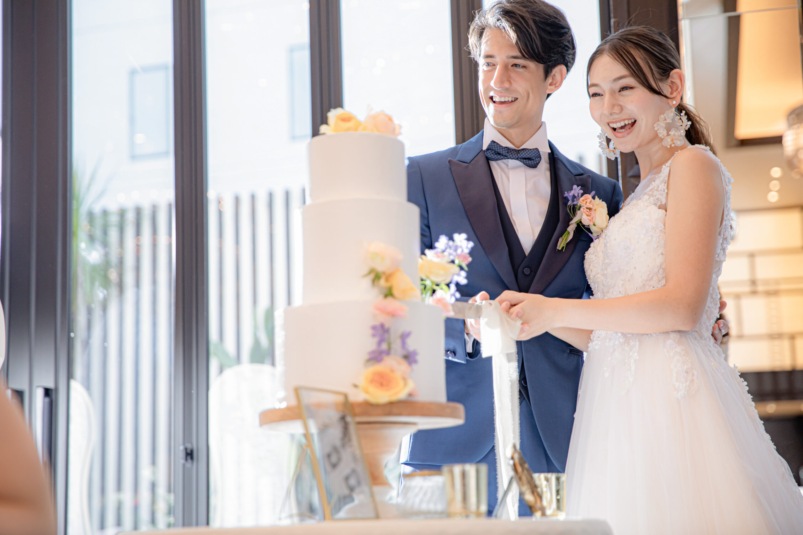 名古屋市の結婚式場ビアンカーラヒルサイドテラスの光溢れる披露宴会場で、笑顔でケーキ入刀を行う新郎新婦。モダンで洗練されたウエディングのワンシーン。