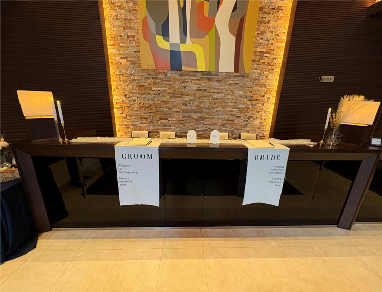 結婚式の受付カウンター。中央にウェルカムサインや芳名帳が並び、左側に“GROOM”、右側に“BRIDE”と書かれた布のタペストリーが垂れ下がっている。背後の壁はストーン調で、カラフルな抽象画が掛けられている。キャンドルやランプが左右に置かれ、落ち着いた雰囲気の装飾が施されている。