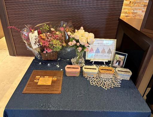 結婚式のウェルカムスペースとして装飾された小さなテーブル。濃紺のクロスの上に花束や花瓶の生花、3色（ブルー・ゴールド・ブラック）の小さなボックス、メッセージを書くボード、写真立てが並ぶ。ライトの装飾もあり、あたたかい雰囲気のディスプレイ。