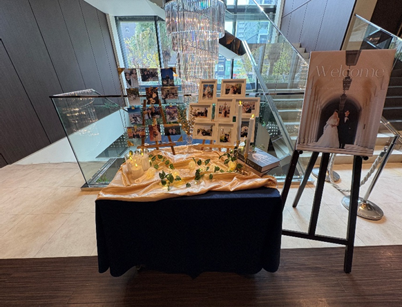 階段前に設置された結婚式のウェルカムスペース。テーブルにはゴールドの布が敷かれ、キャンドルやグリーンの装飾、ふたりの写真をまとめたフレームが並んでいる。左奥には多数の写真が吊るされたフォトディスプレイ、右側には新郎新婦のウェルカムボードがイーゼルに立てられている。