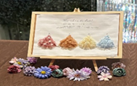 ブラウンのテーブル上に、ピンク・オレンジ・イエロー・ブルーの4色のフラワーアートを額に入れたウェルカムボードと、カラフルな造花が並べられた結婚式の受付ディスプレイ。