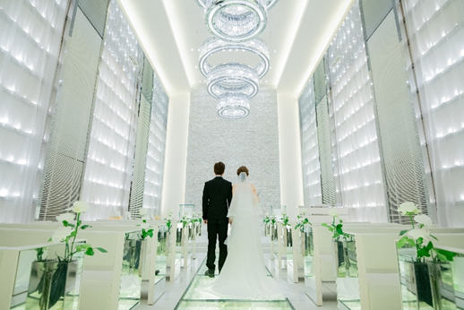 クリスタルが輝くガラスのバージンロードに立つ新郎新婦の後ろ姿。5連のリング型シャンデリアと白い大理石の壁が美しい、ビアンカーラ・ヒルサイドテラスの幻想的なチャペル。