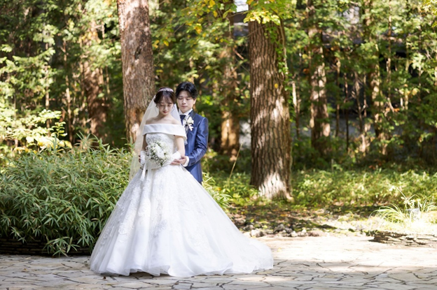 名古屋市の結婚式場ビアンカーラヒルサイドテラスが贈る森のロケーションフォト。木漏れ日溢れるガーデンで、オフショルダーのドレスを纏った新婦と新郎が描く幻想的な一枚。