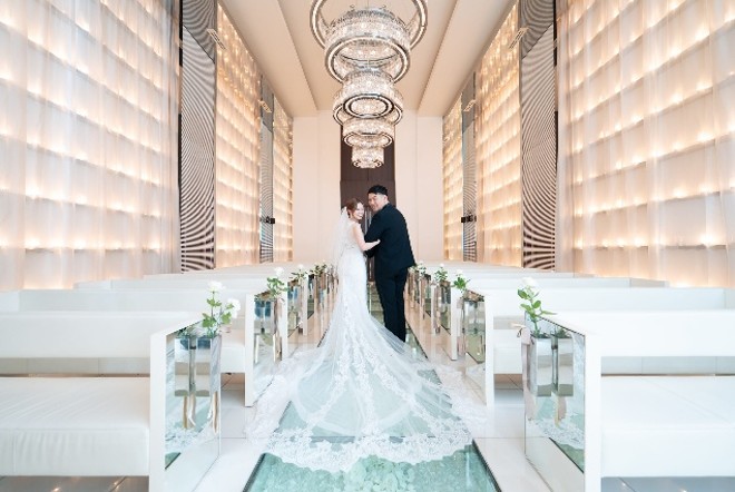 名古屋市の結婚式場ビアンカーラヒルサイドテラス。光り輝くモダンなチャペルで、長いトレーンのウェディングドレスを着た新郎新婦が微笑む様子。